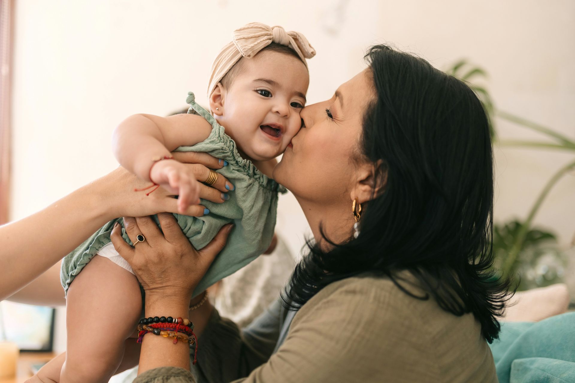 Woman kisses a baby held up by another person. Baby has a happy expression.