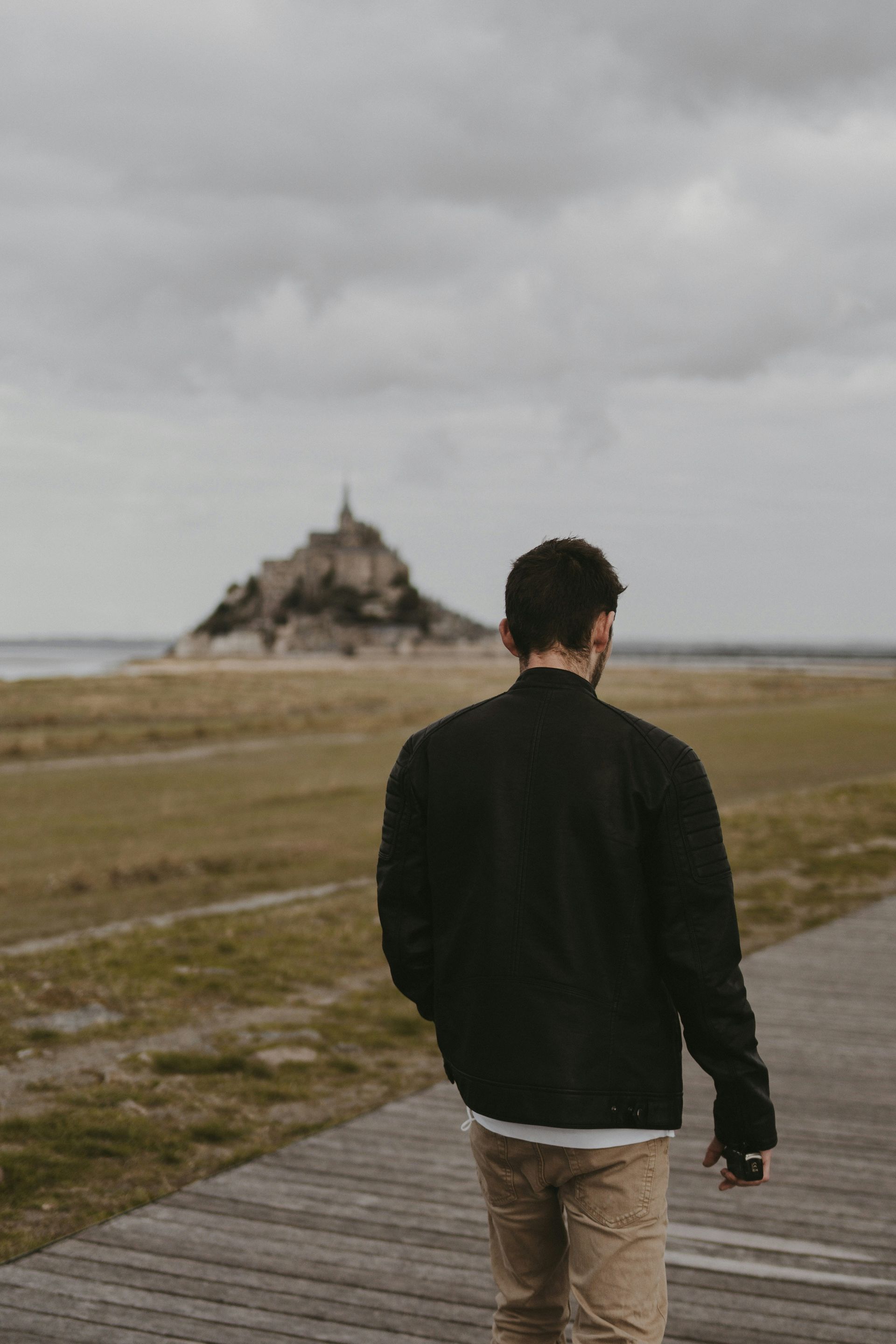 Man walks on a path, facing away from camera, towards Mont Saint-Michel under overcast skies.
