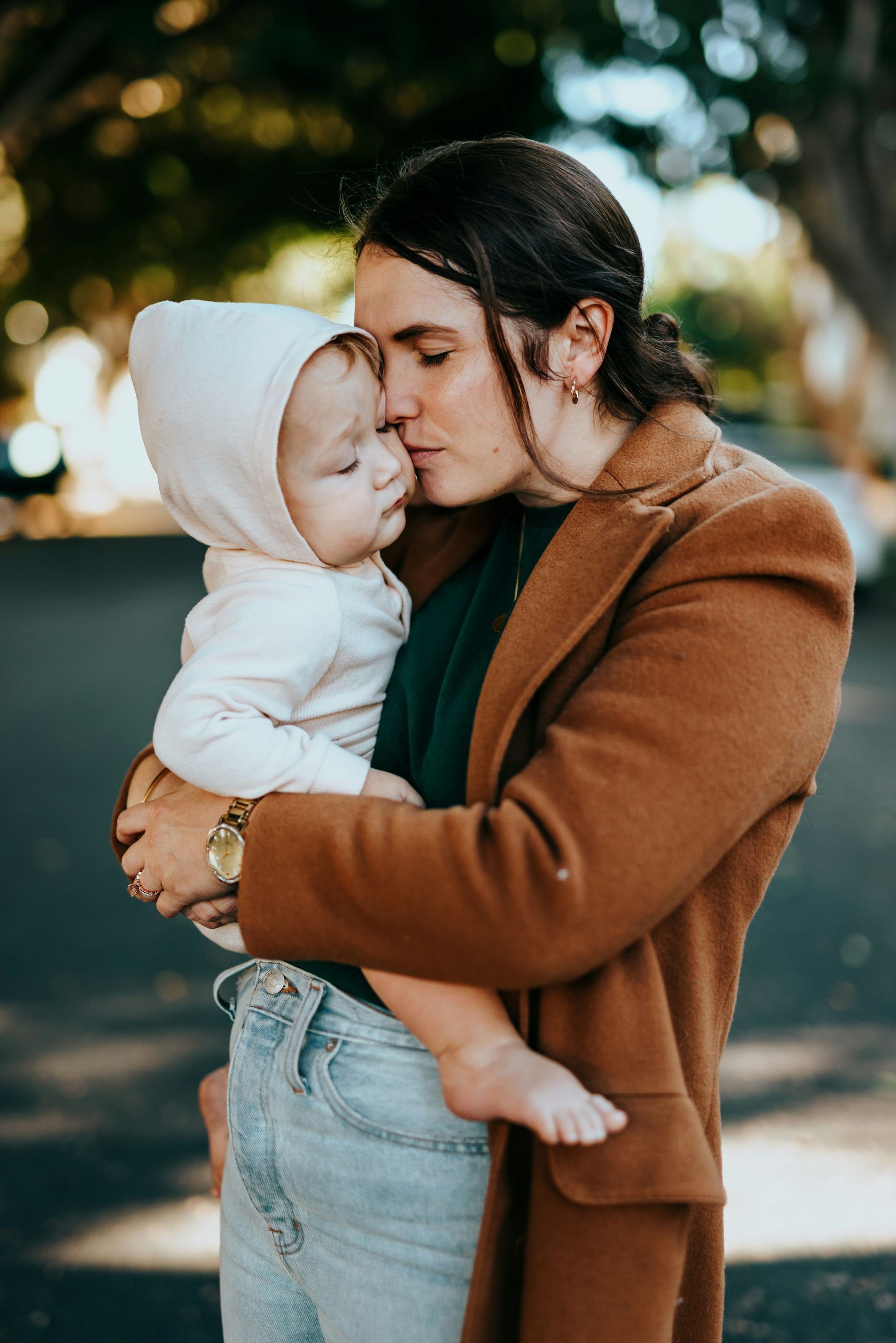 Woman in brown coat kisses baby in white hooded onesie on cheek outdoors.