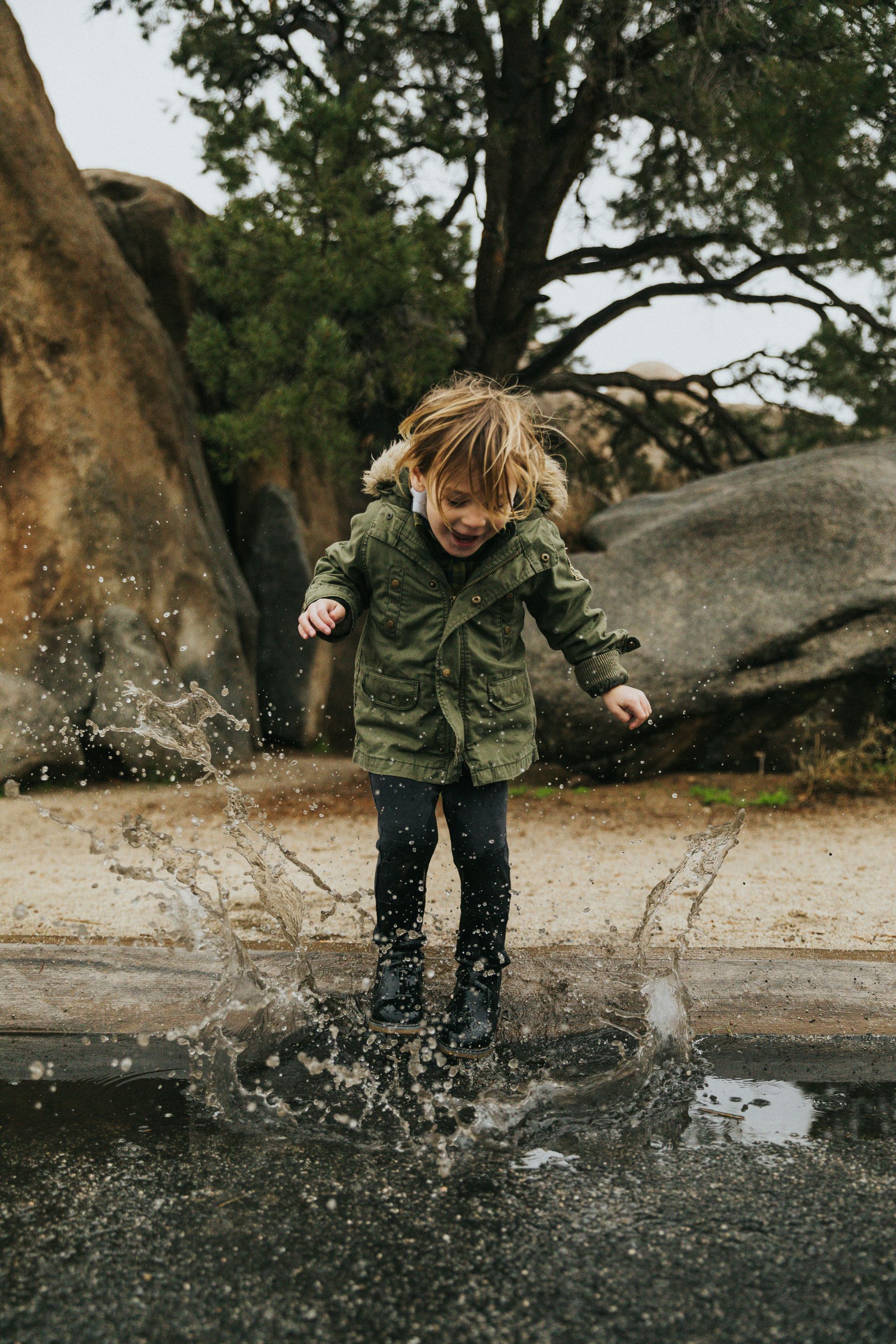 Boy in green jacket jumps in puddle, splashing water. Brown rocks and trees in the background.