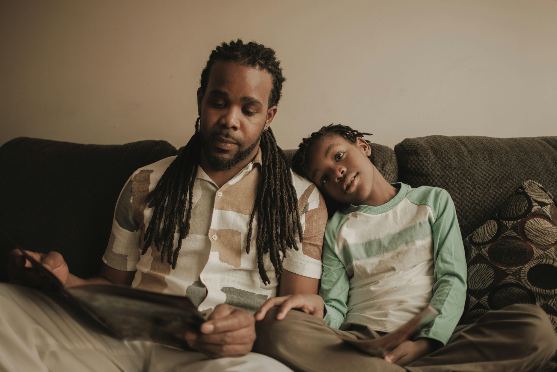 Father with dreadlocks reads to his son on a couch. Son rests head on dad’s shoulder, both focused.