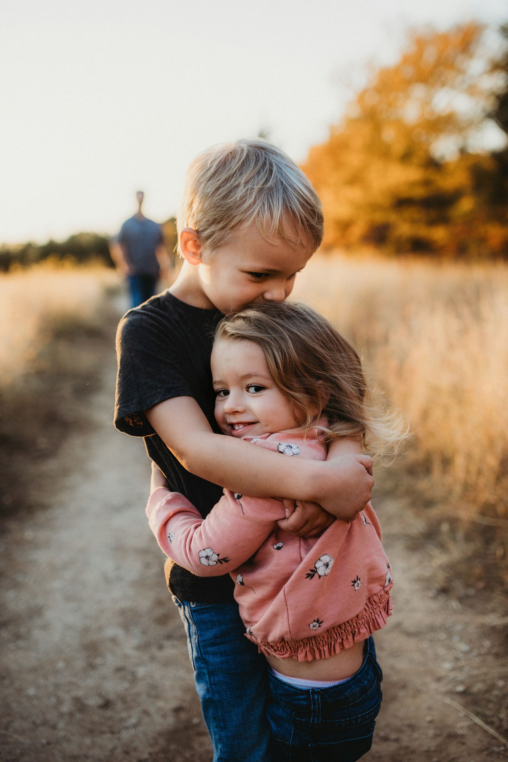 Blonde boy hugging and kissing a smiling girl; dirt path, golden hour. A man walks in the background.