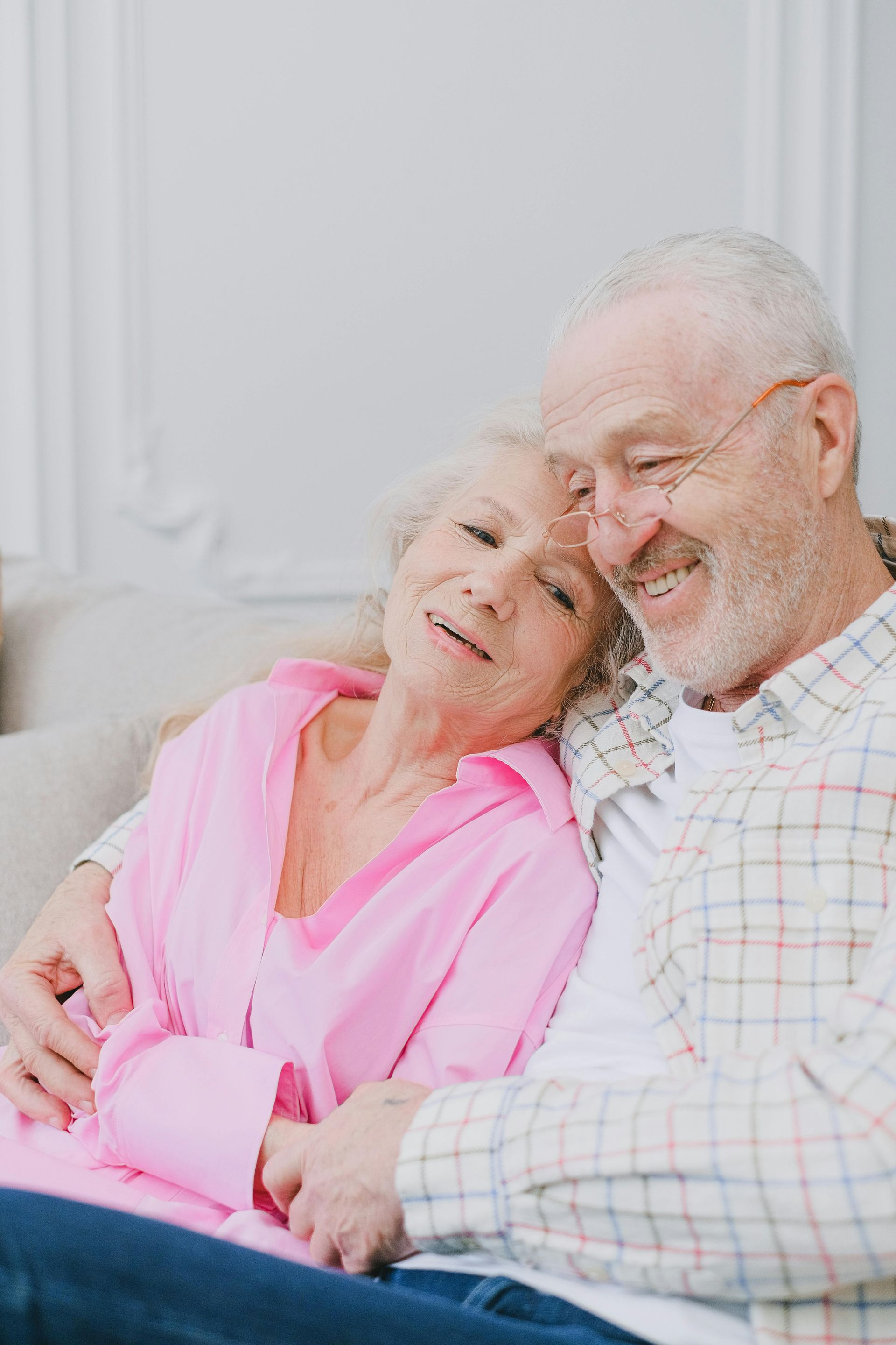 Elderly couple embraces on a couch; woman in pink, man in plaid, both smiling warmly.
