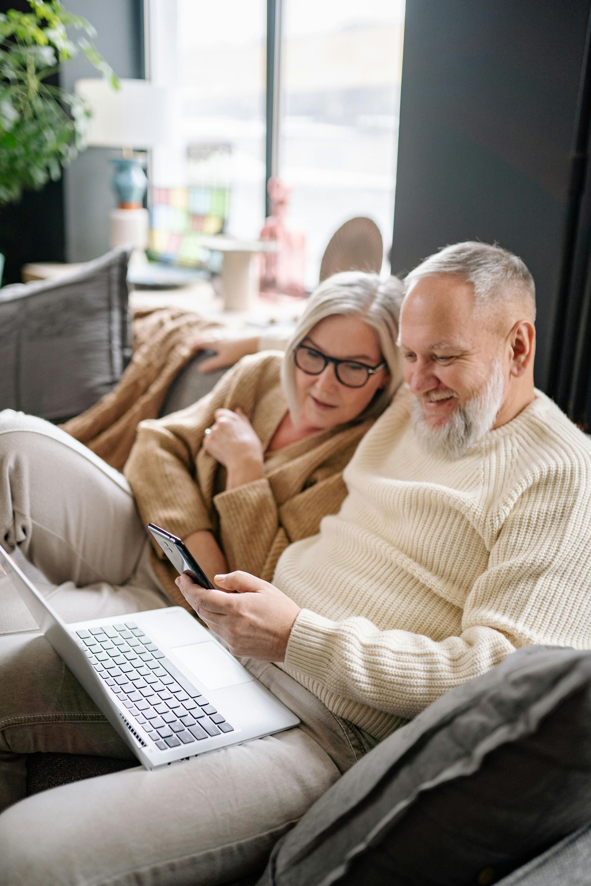 Senior couple on sofa looking at a laptop and phone, smiling. Inside living room.