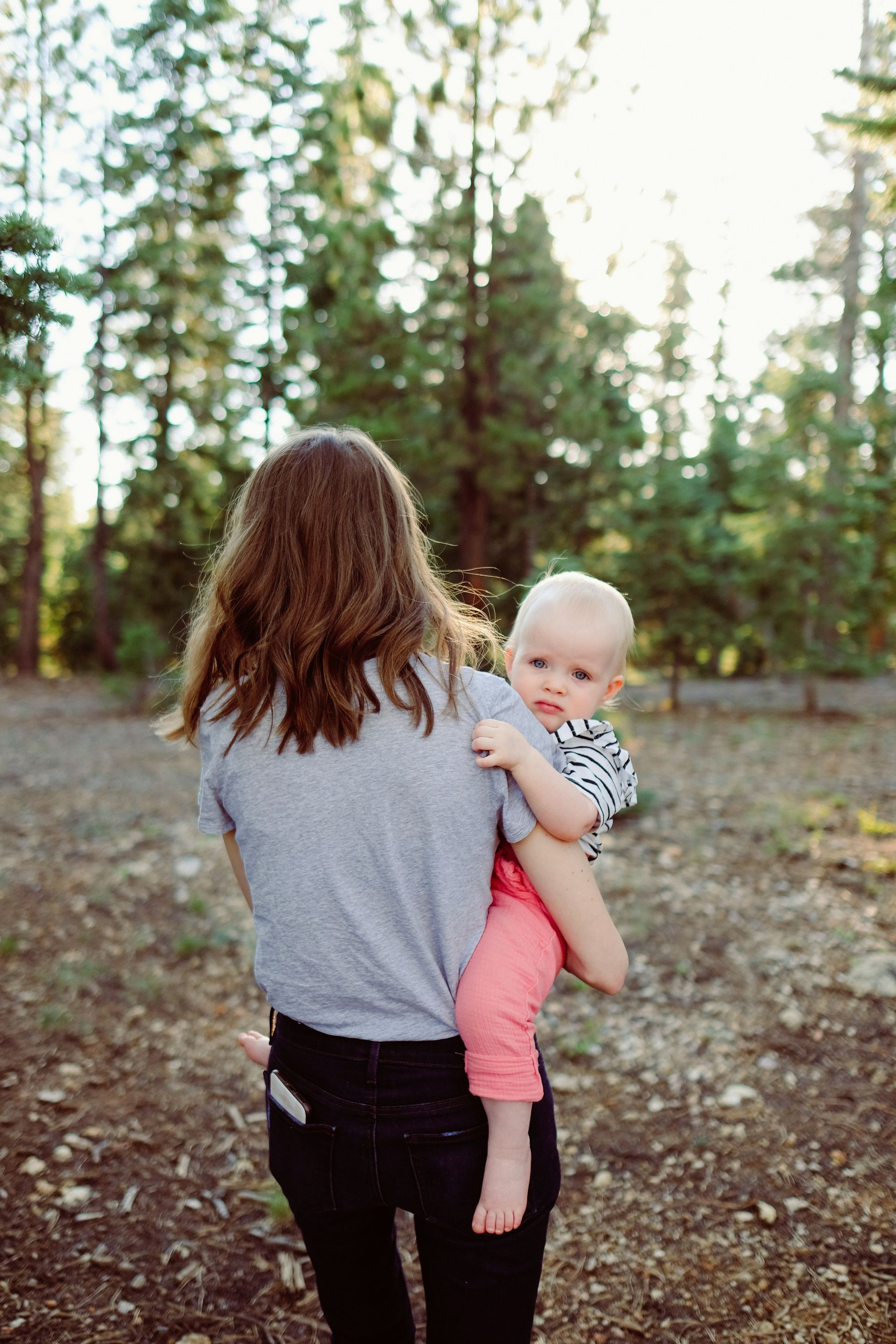Woman holding baby in forest. Baby looks at camera, wearing pink pants and striped shirt.
