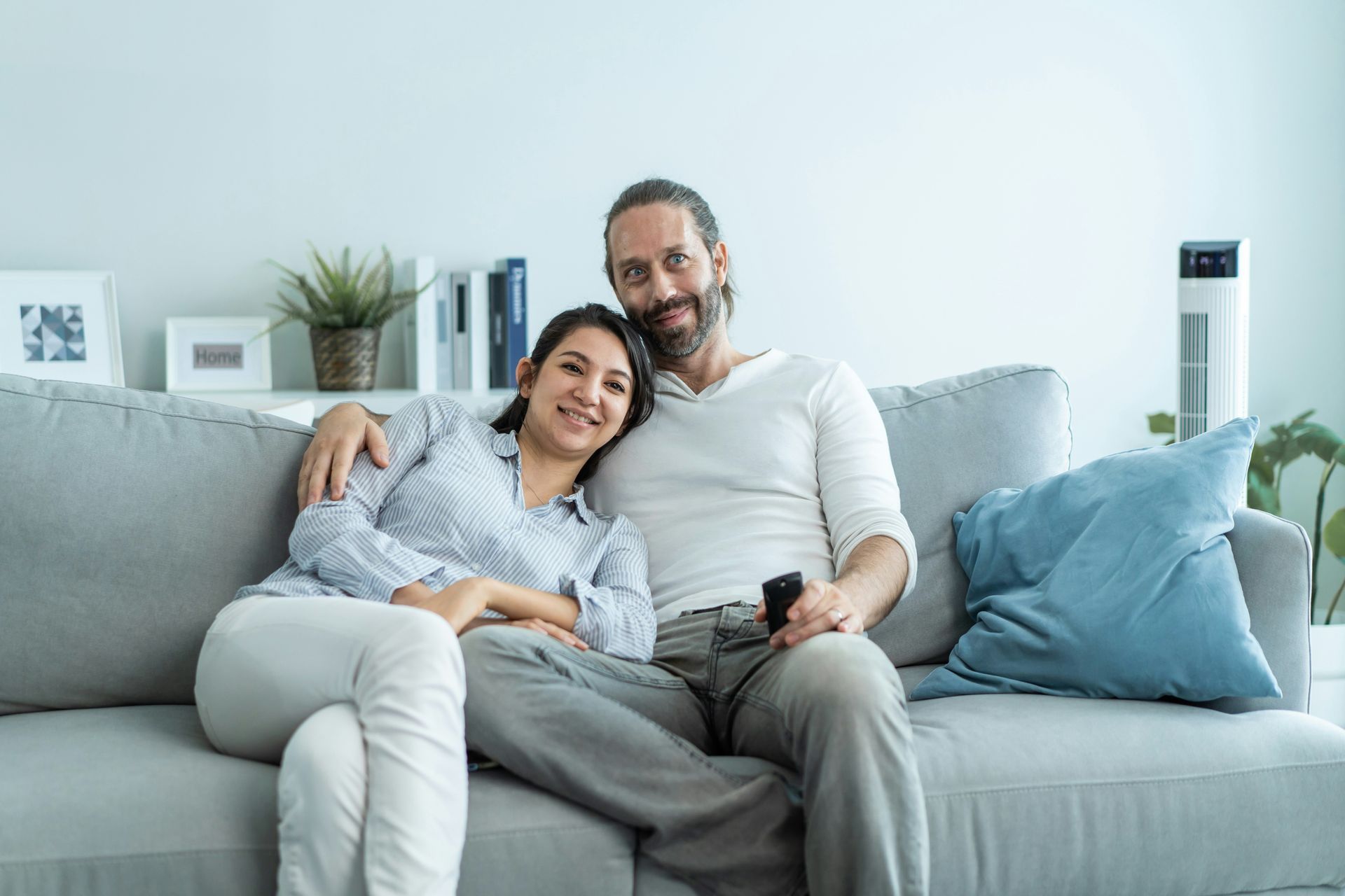Couple cuddling on a gray sofa, smiling at the camera. They are inside a bright living room.