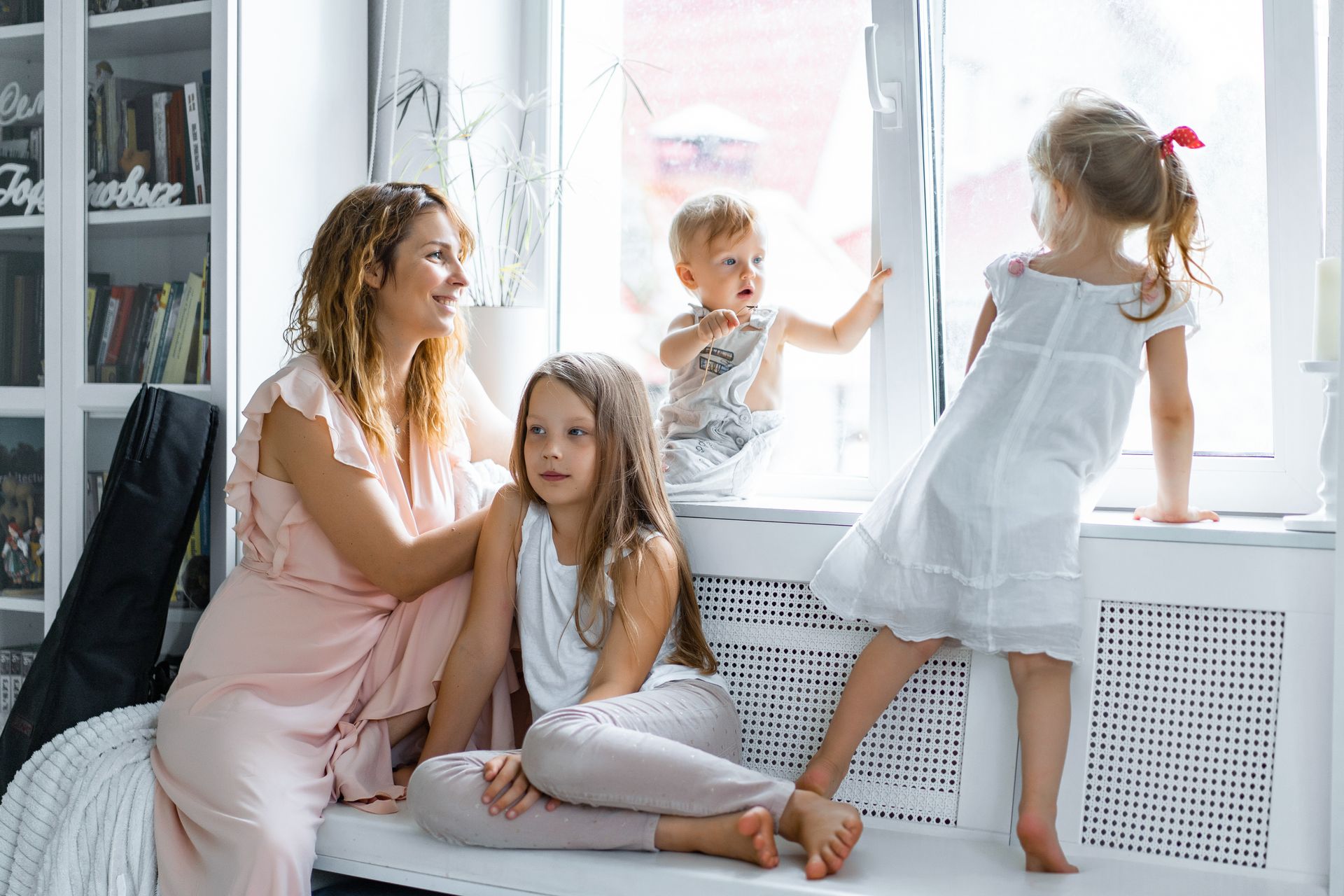 Woman and three children by a window, one looking out, one on lap, all smiling.