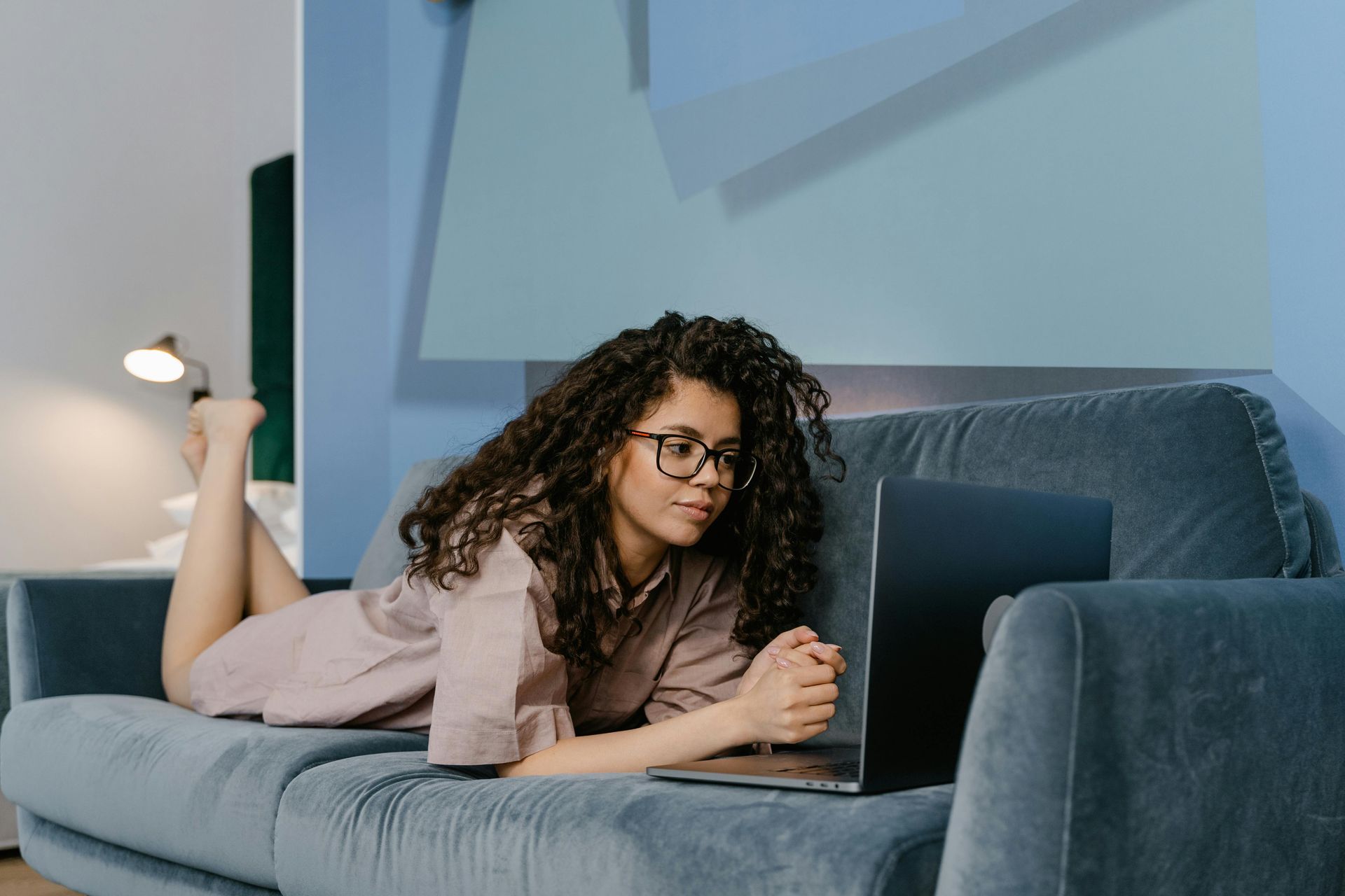 Woman with curly hair and glasses lying on a blue couch, working on a laptop.
