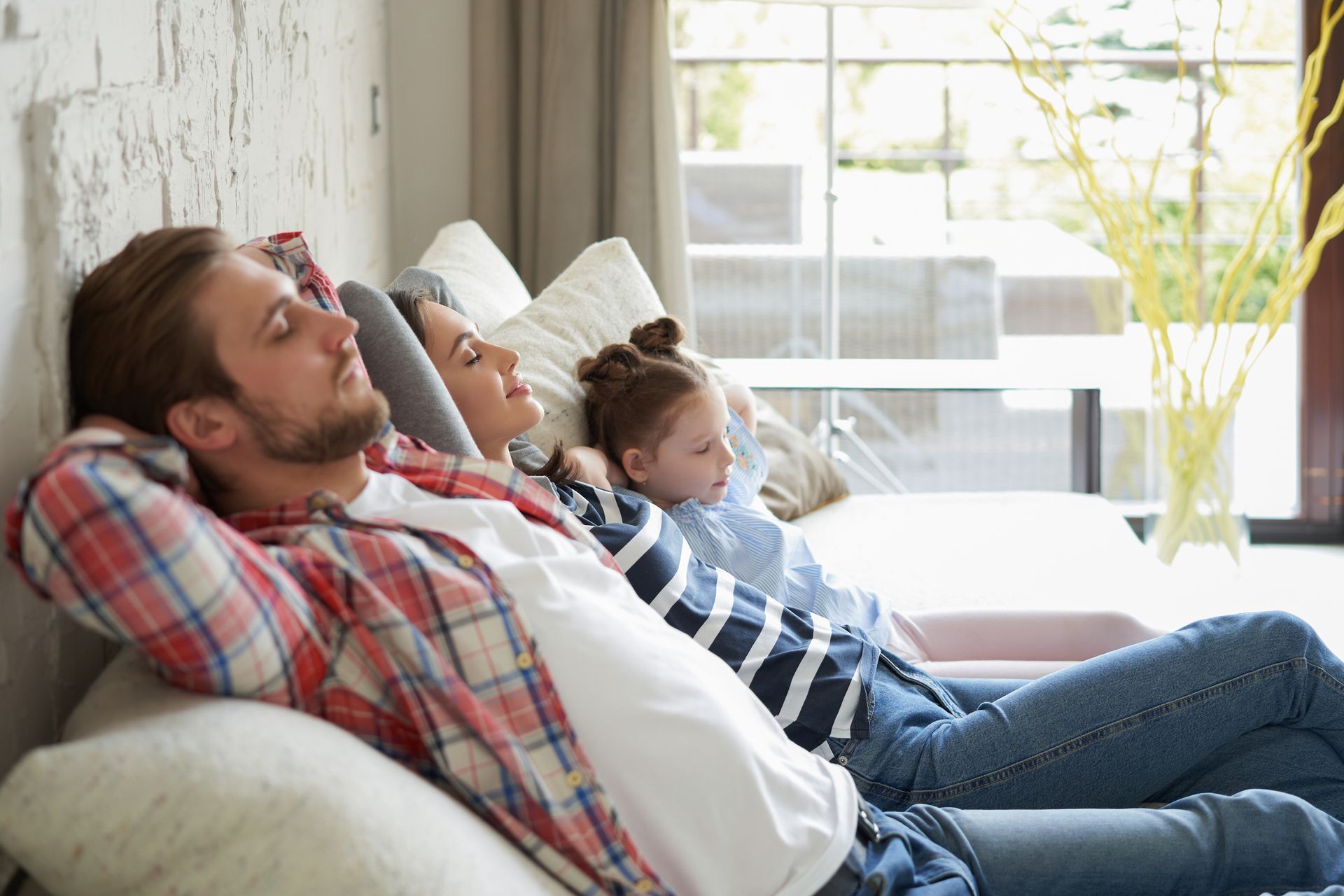 Family of three resting on a couch; man with hands behind head, woman and child cuddling, all eyes closed.