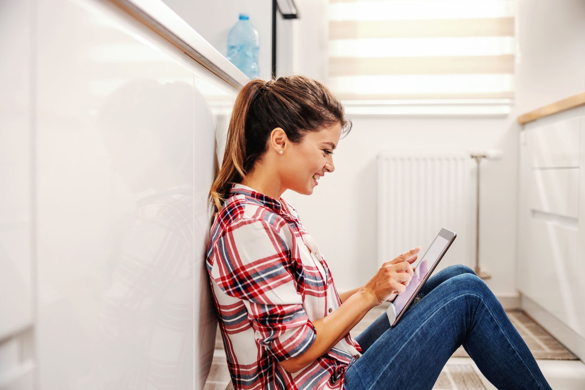 Woman in plaid shirt and jeans sits on floor, using a tablet near white cabinets.