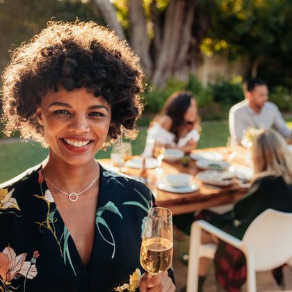Smiling african woman standing outdoors with people sitting in background having food at party. Woman with a drinks at outdoor party.