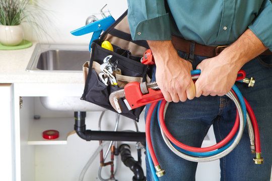 A plumber is holding a wrench and a hose in front of a sink.