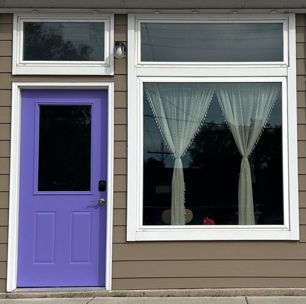 Purple door and large window with white curtains on a brown building.