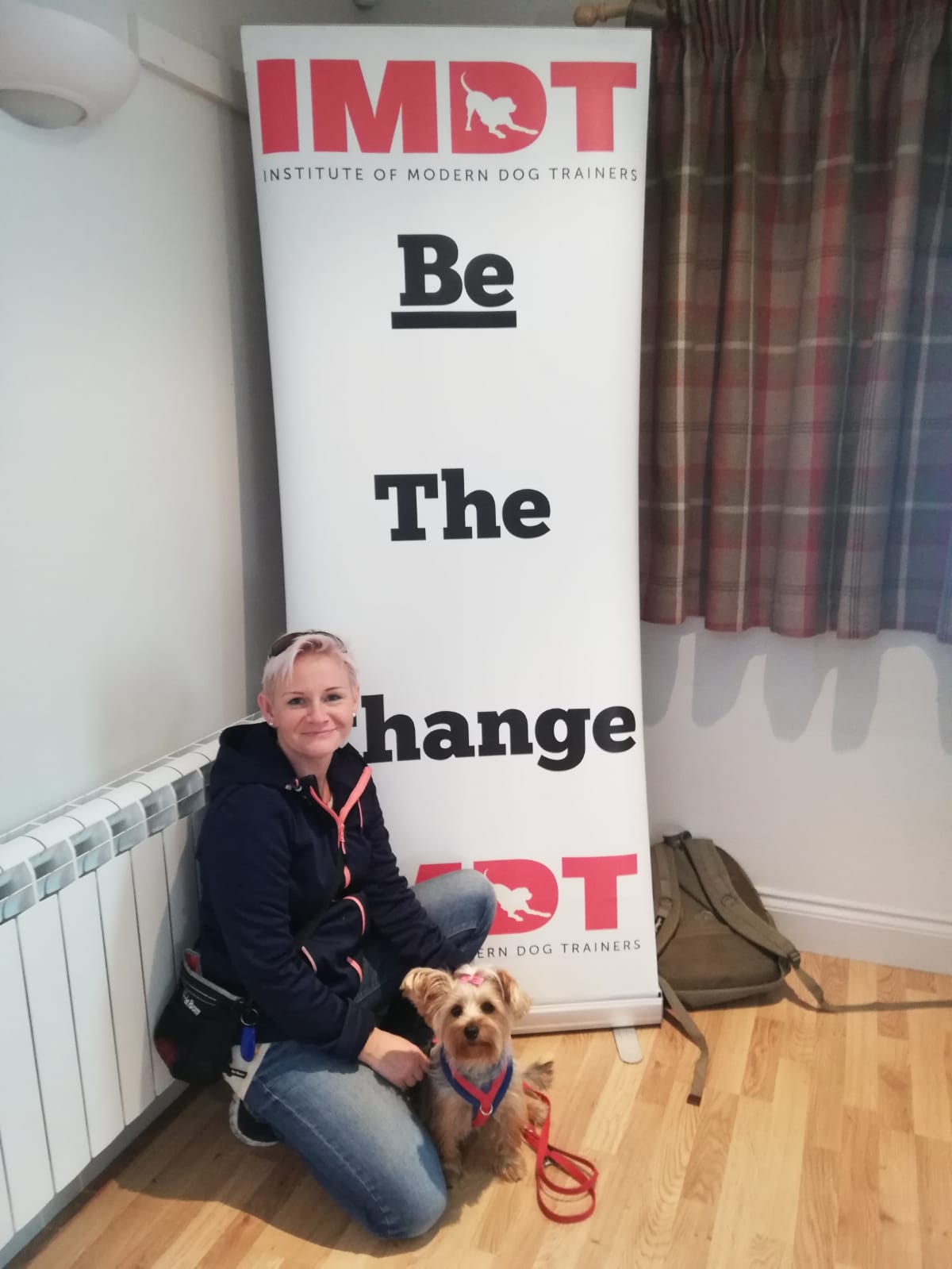 Middle-aged woman knelt with her dog in front of a banner for the Institute of Modern Dog Trainers