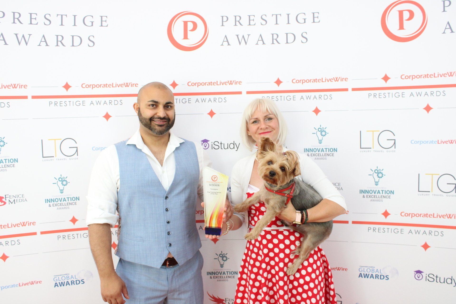 A woman in a spotty dress holding her small dog under one arm and a dog service provider award in the other hand.