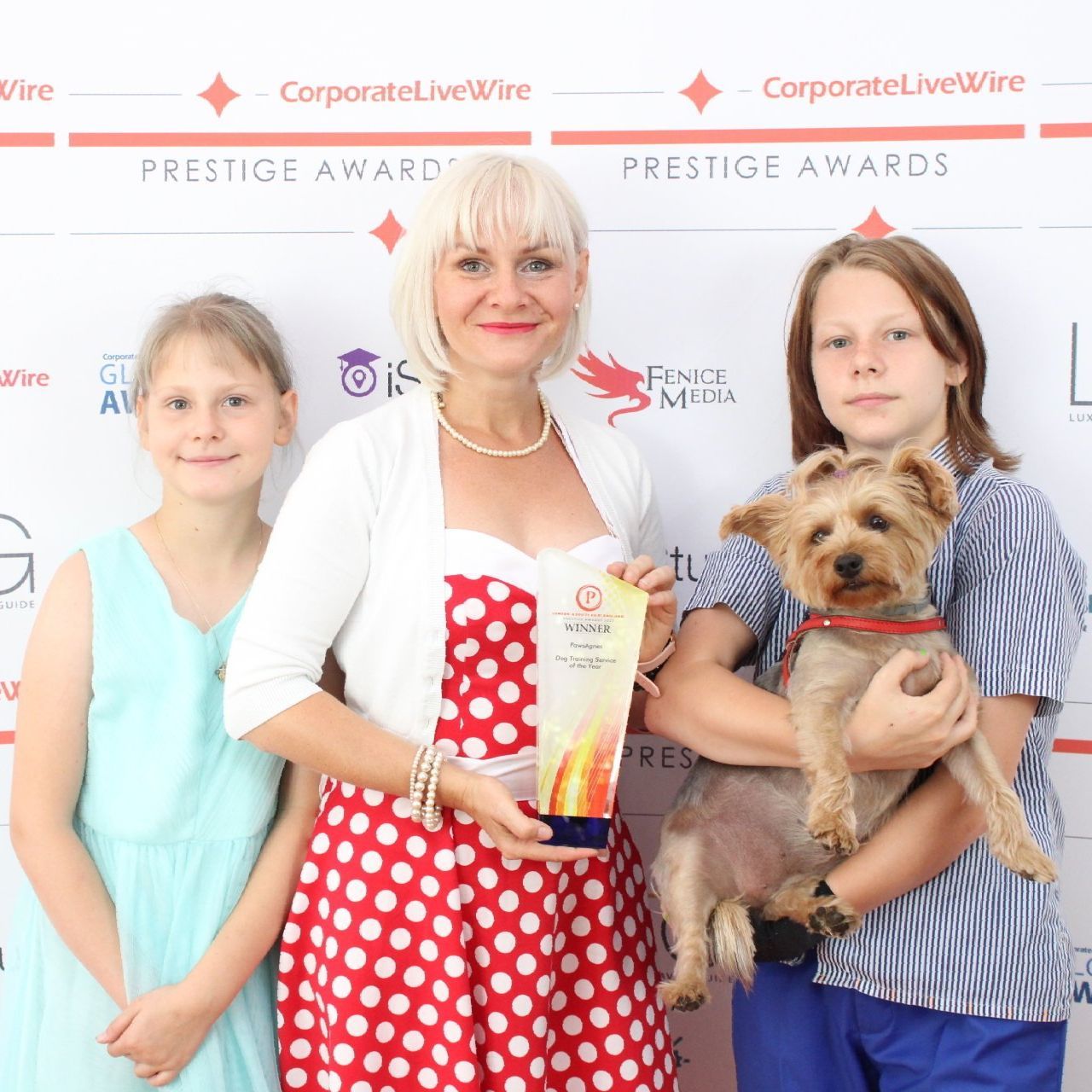 A middle-aged woman in a spotted dress holding an award. Two children stand either side, one holding a small dog.