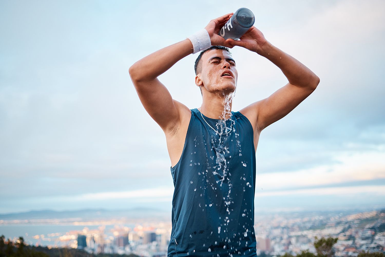 A person in a blue tank top pours water over their face to cool down, with a blurred city skyline in the background.