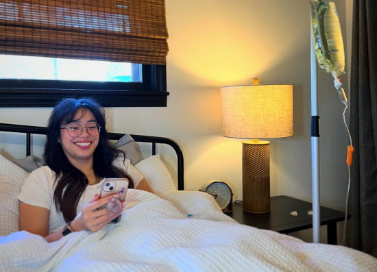 A woman is laying in bed with an iv in her arm and smiling while looking at her phone.