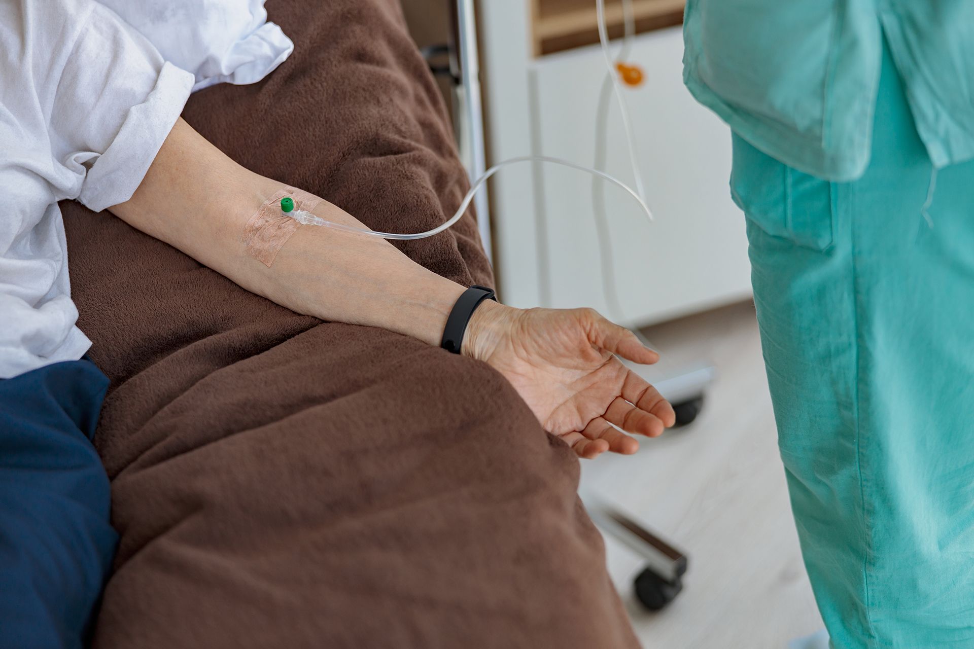 Patient arm with IV line, seated beside a healthcare worker in green scrubs in a clinic room