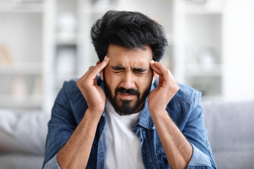 A man with a beard is sitting on a couch with a headache.