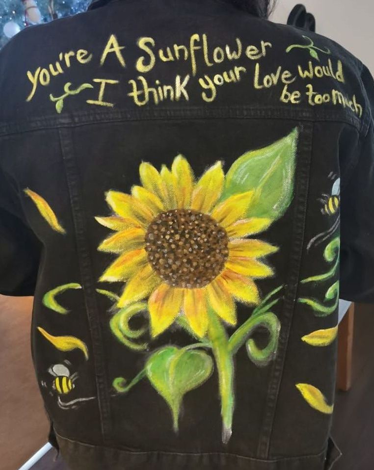 A sunflower is painted on the back of a black jacket, with buzzing bees and floating petals.  The words 