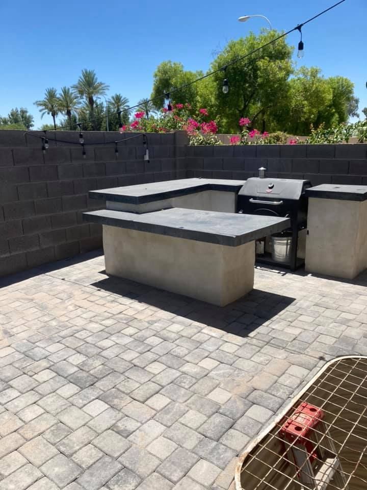 Outdoor kitchen with concrete countertops, a grill, and a brick patio on a sunny day.