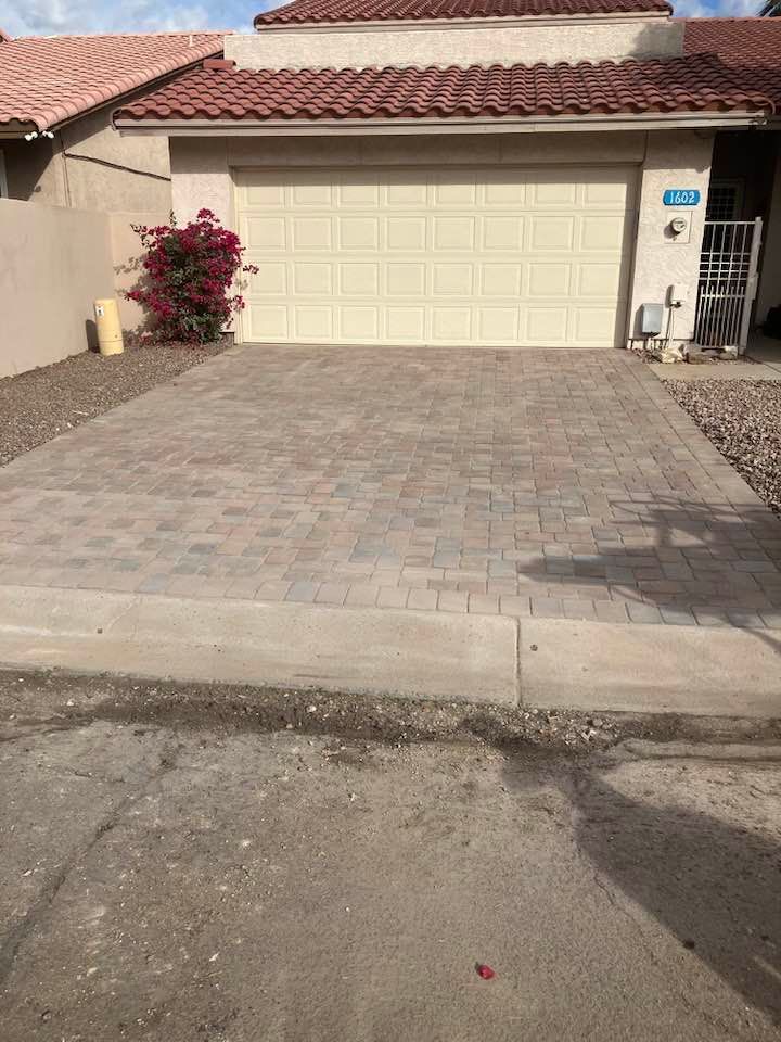 Brick paved driveway in front of a house with garage; small red bush to the left.