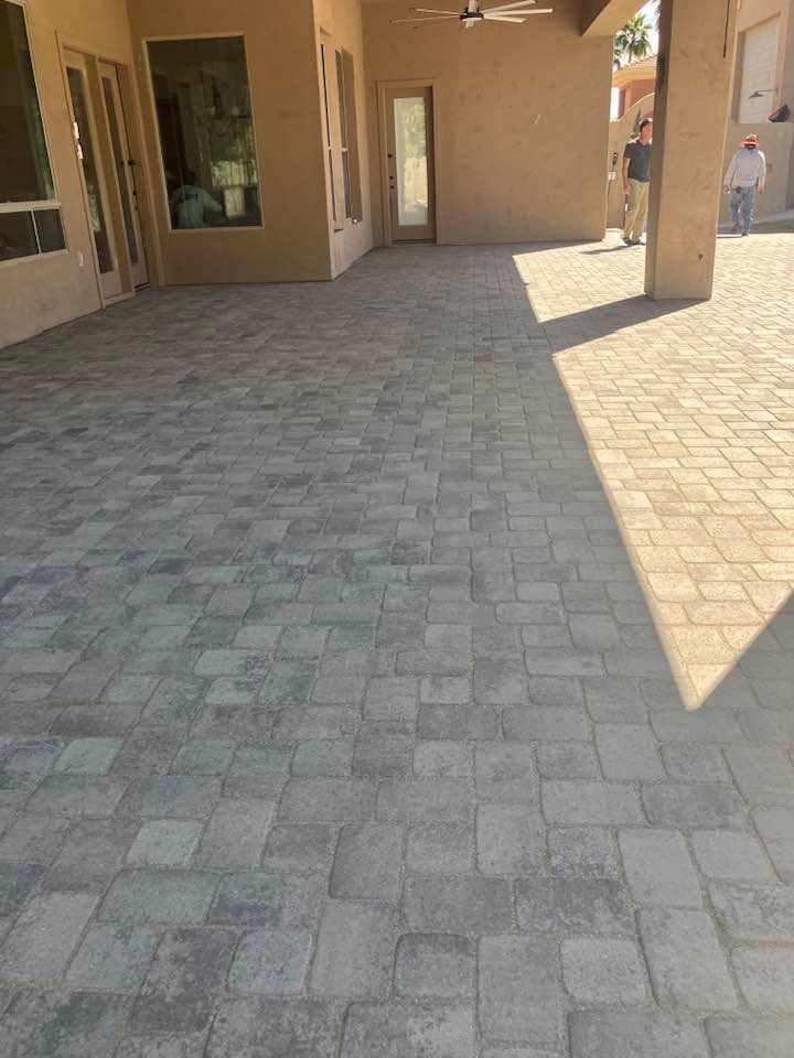 Patio paved with gray bricks, under an overhang, with sunlight, and two people in the background.