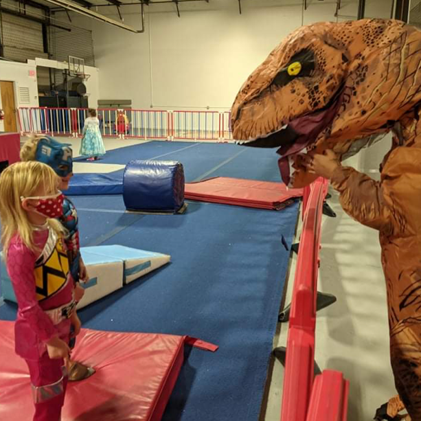 A little girl wearing a mask stands next to a dinosaur costume