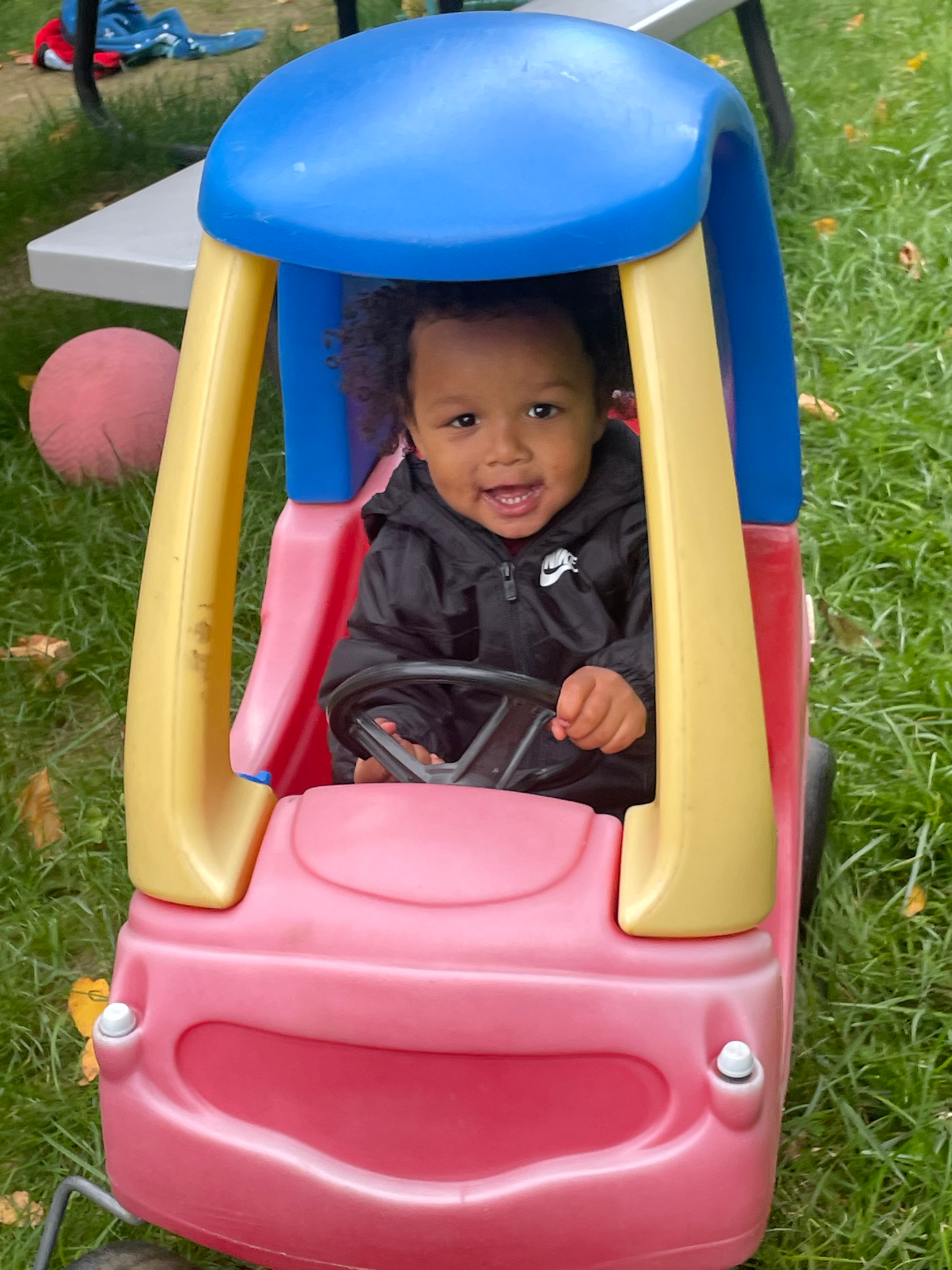 A baby is sitting in a pink toy car in the grass.