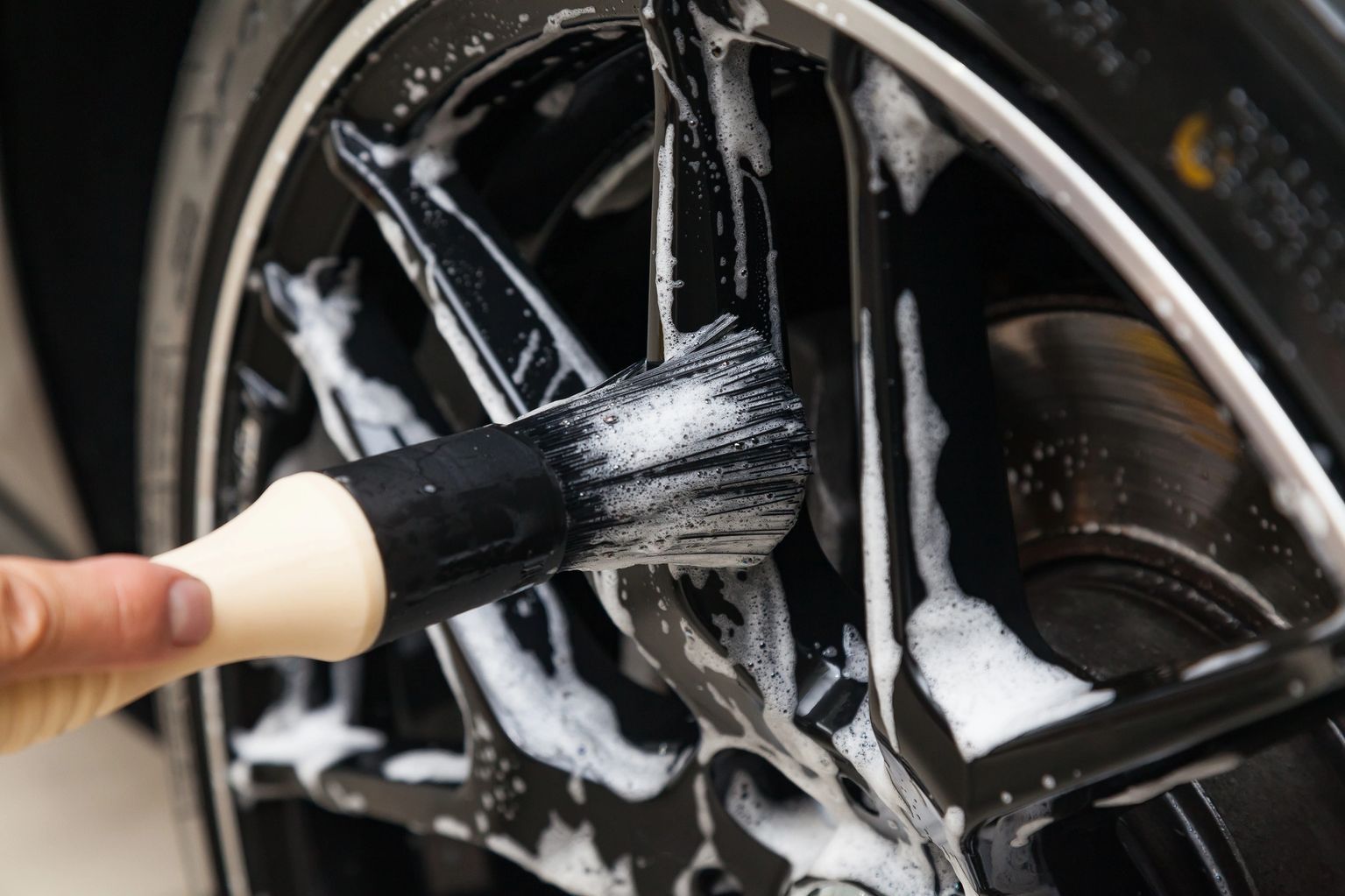 A hand brushes soapy suds onto a black car wheel rim during cleaning.