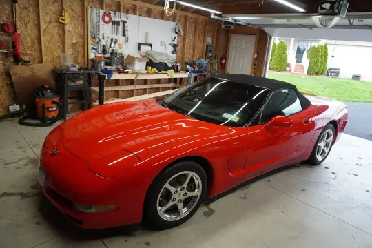 Red sports car parked in a cluttered garage with the door open to a driveway.