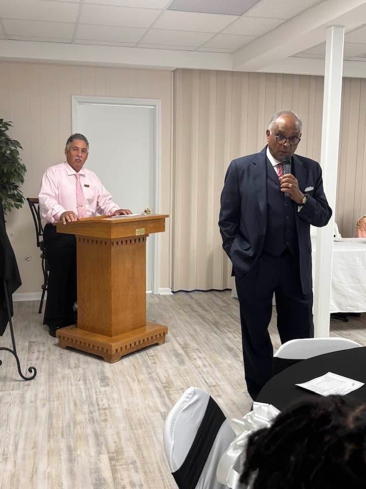 A man in a suit is giving a speech at a podium while another man sits at a table.