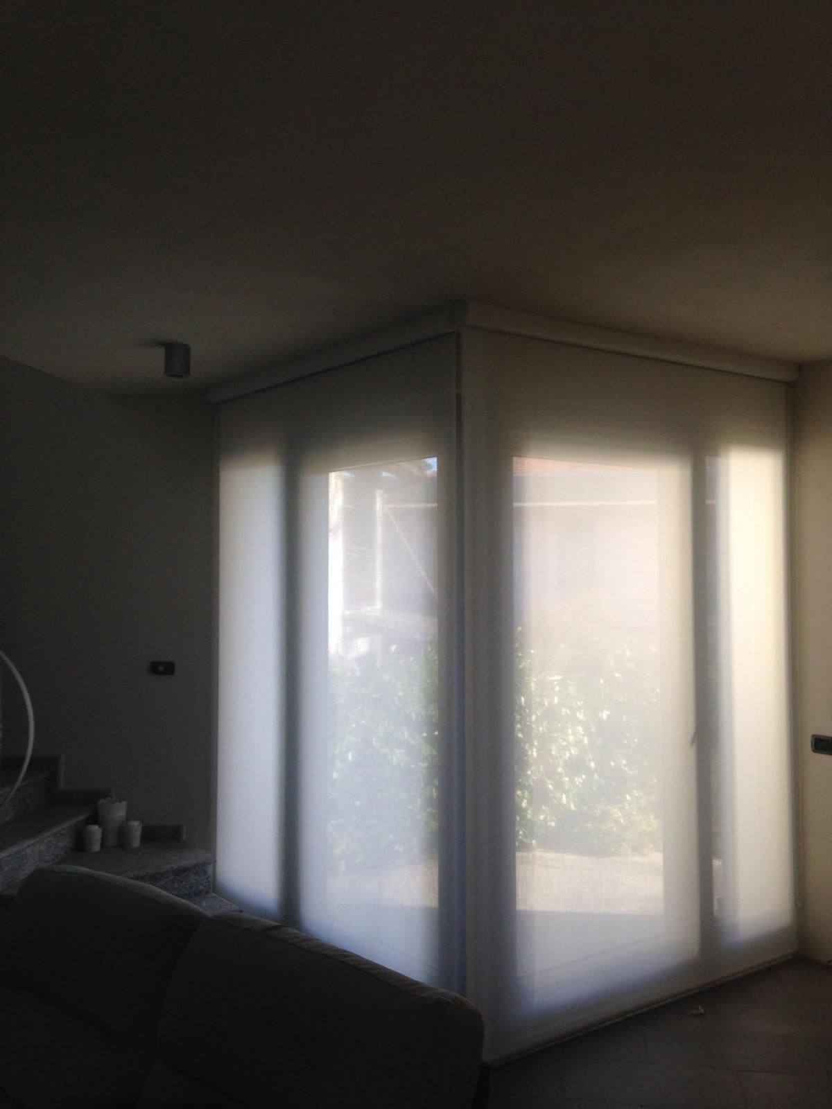 Ampie finestre a tutta altezza, coperte da tende a rullo bianche traslucide, nell'angolo di un soggiorno.