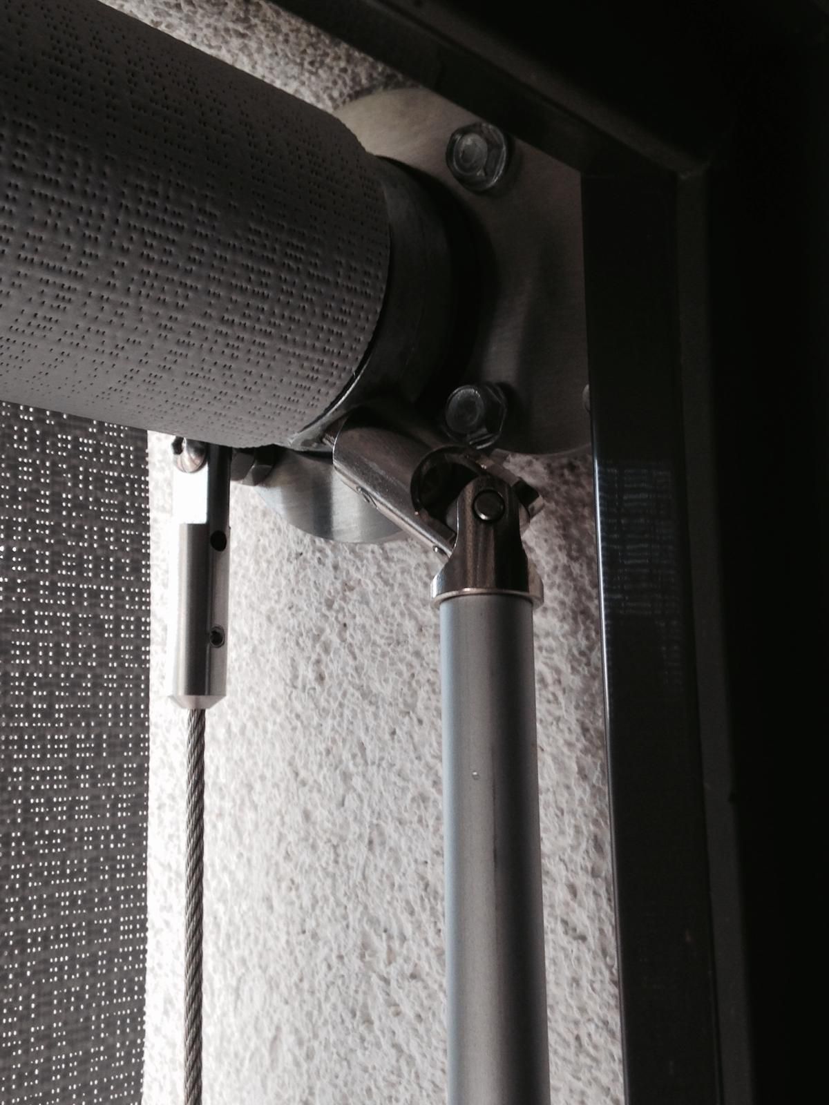 Primo piano di un meccanismo metallico per tende da finestra, che mostra il tubo avvolgitore fissato a una staffa e un'asta di azionamento snodata.
