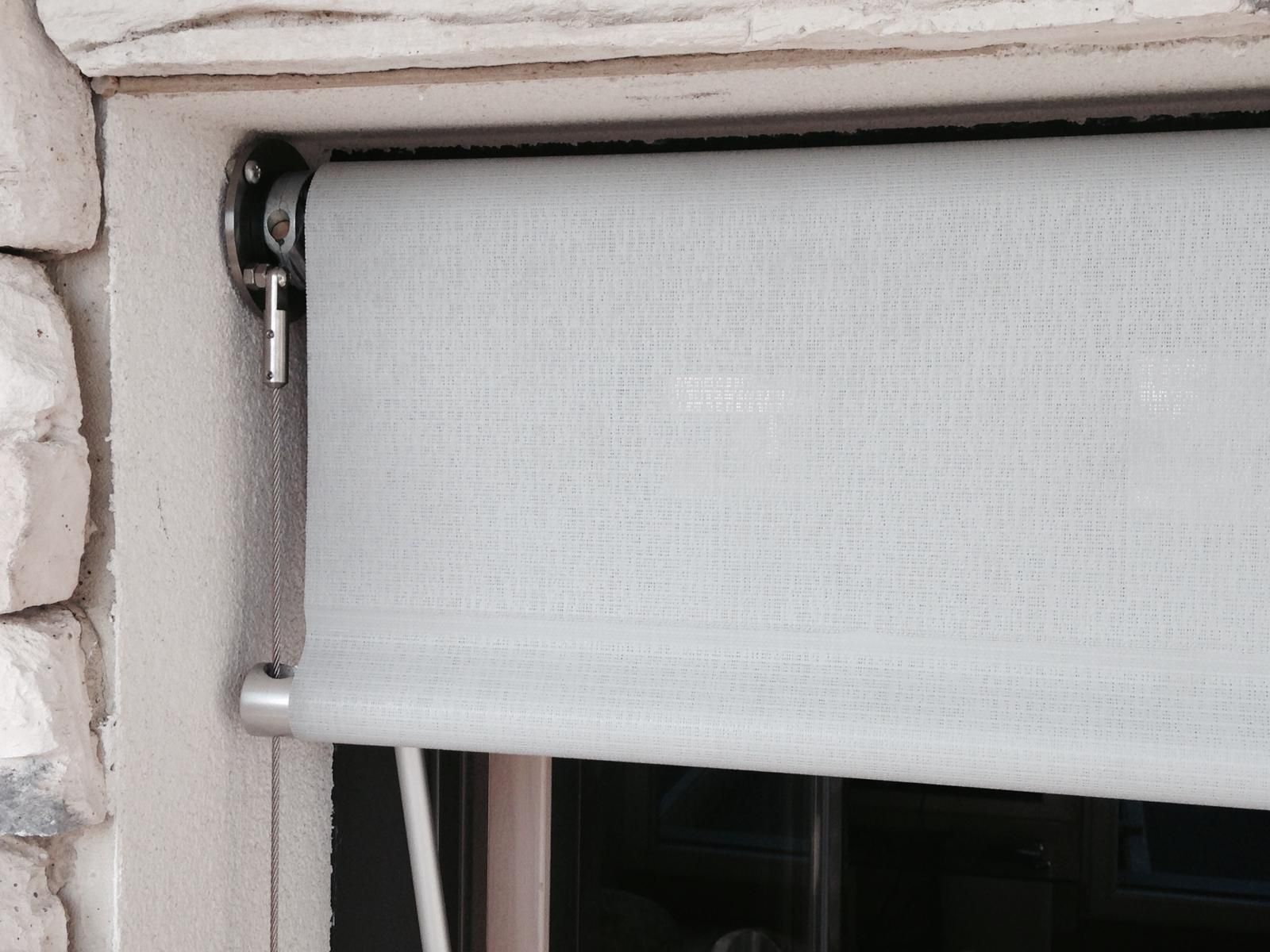 Una tenda a rullo grigio chiaro montata all'interno di un telaio di finestra contro un muro di pietra, che mostra la staffa metallica e la catena.