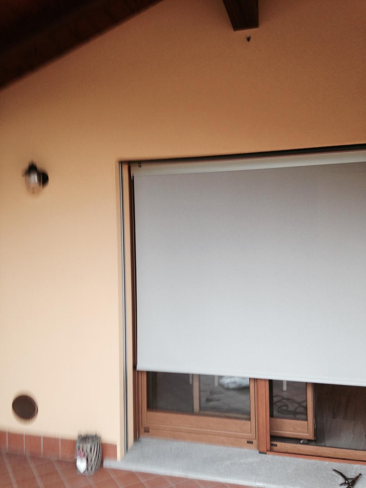 Una parete beige con una lampada a parete, piastrelle in terracotta sul pavimento e una tenda a rullo grigia parzialmente abbassata sopra una porta a vetri.