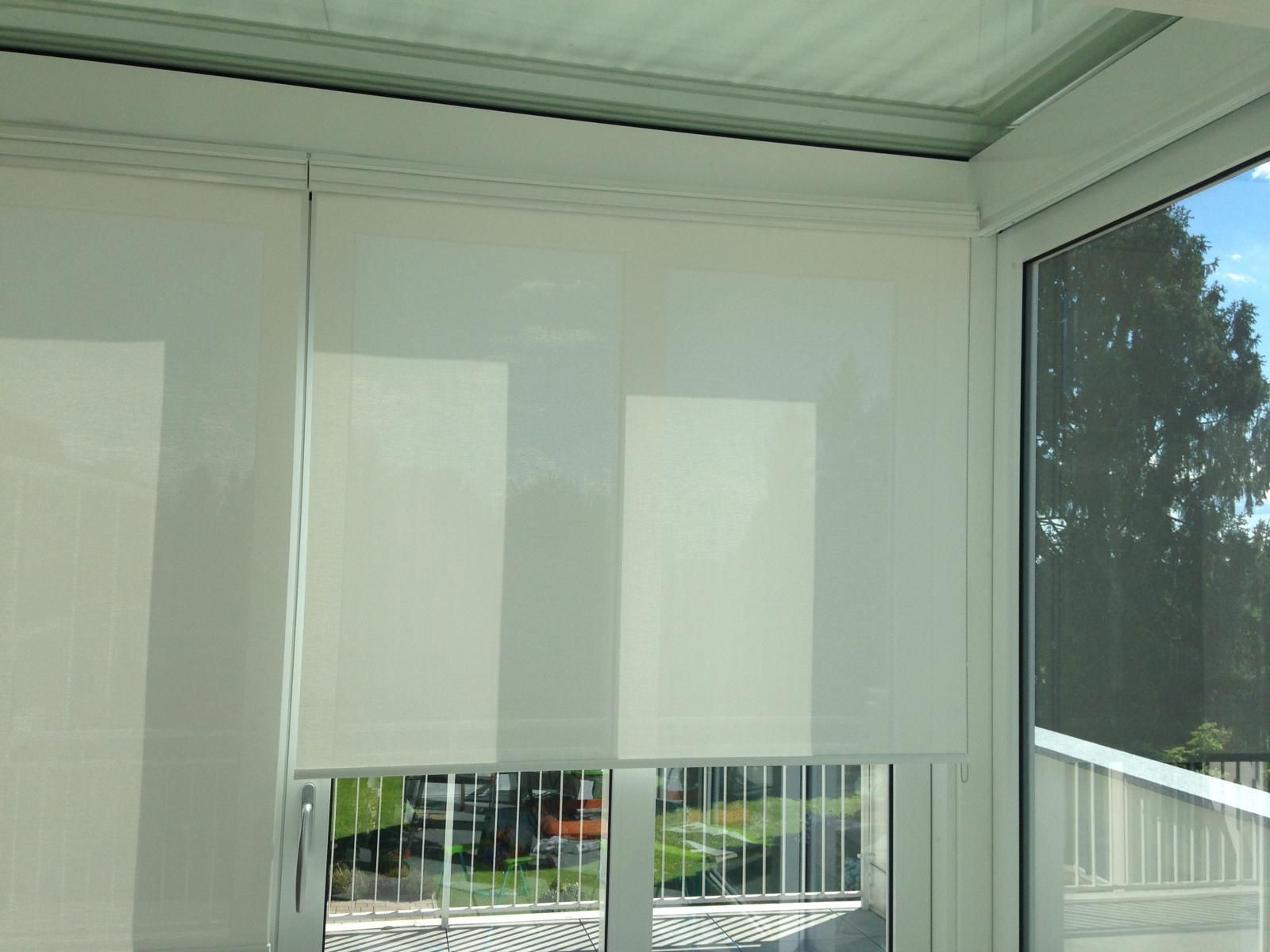 Due tende a rullo bianche montate su un telaio di finestra bianco in una veranda, con vista sugli alberi e sul balcone esterno.
