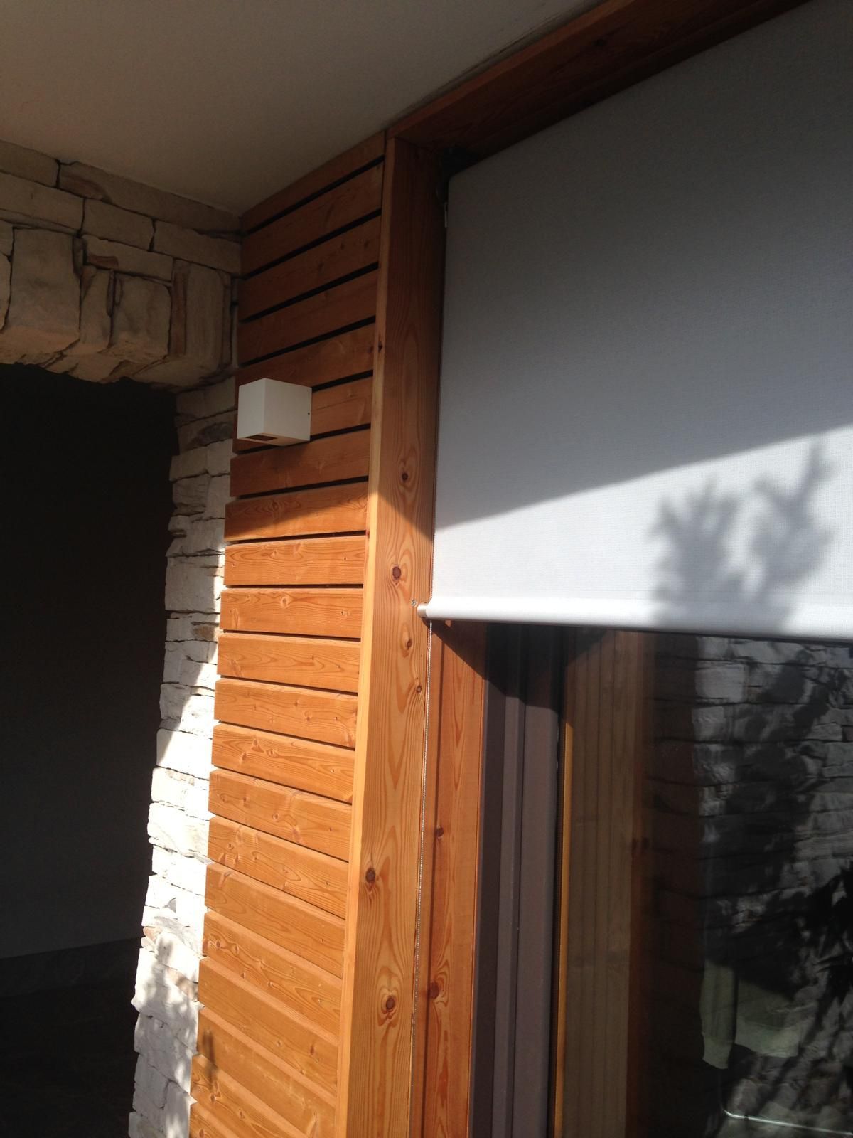 Una tenda bianca parzialmente abbassata accanto a una parete a doghe di legno e a un pilastro di pietra su un patio esterno.