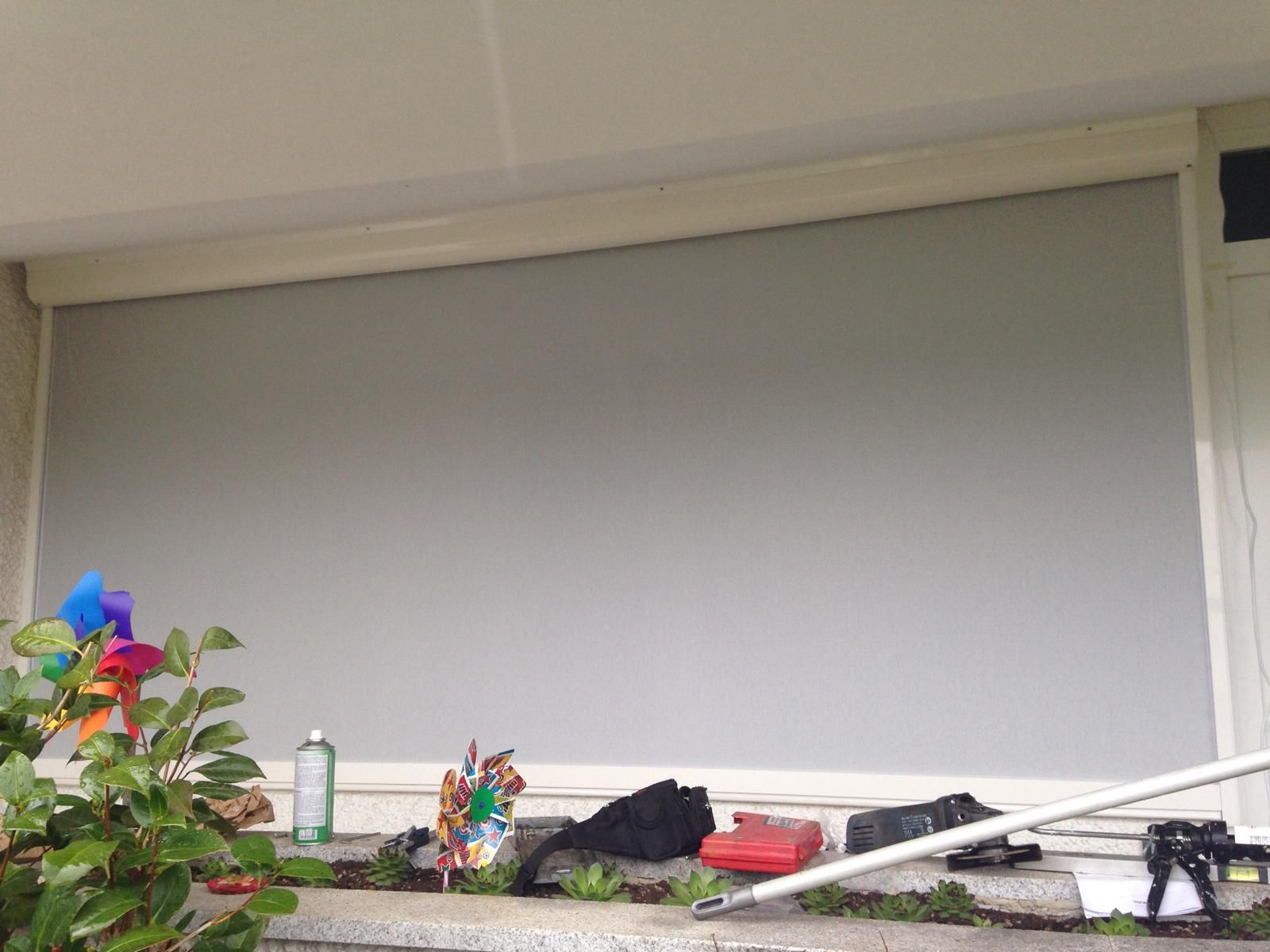 Una grande tenda a rullo da esterno di colore grigio chiaro copre una finestra, sulla quale si trovano una pianta e degli attrezzi.
