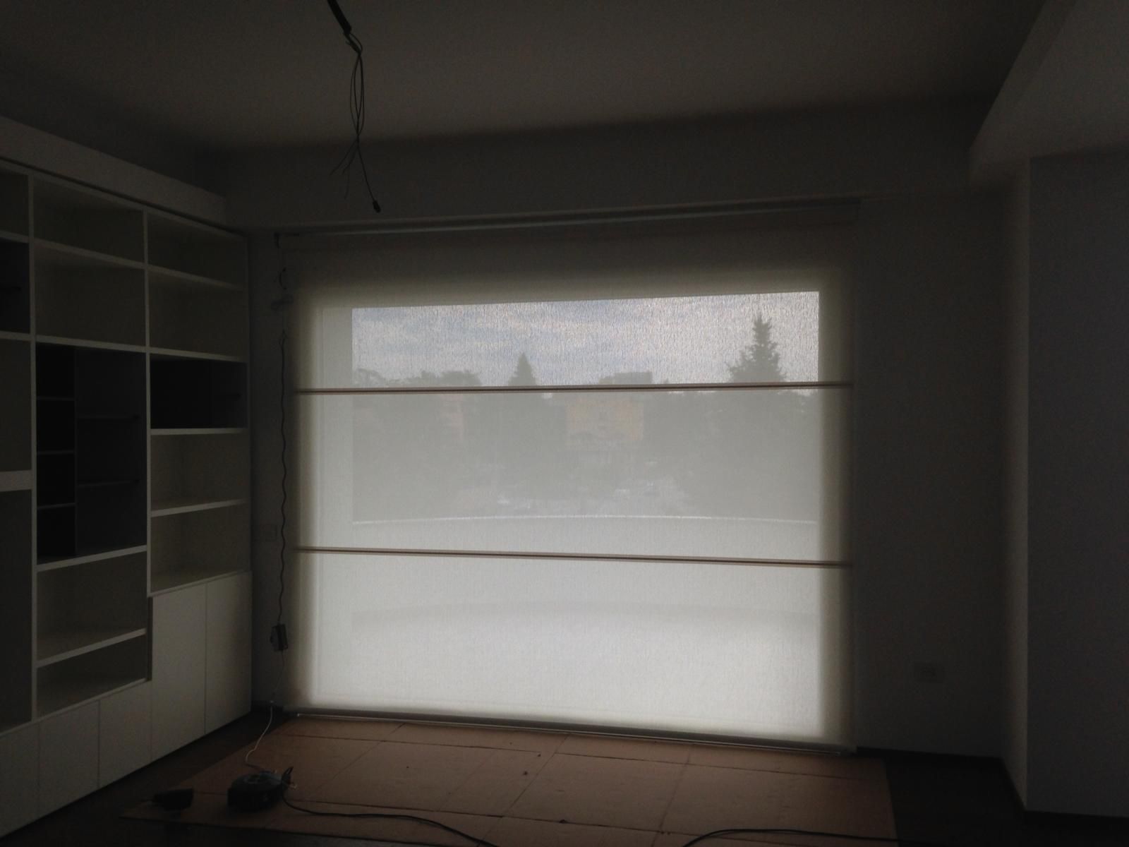 Una grande finestra con una tenda bianca che oscura la vista, accanto a una scaffalatura alta e bianca in una stanza poco illuminata.