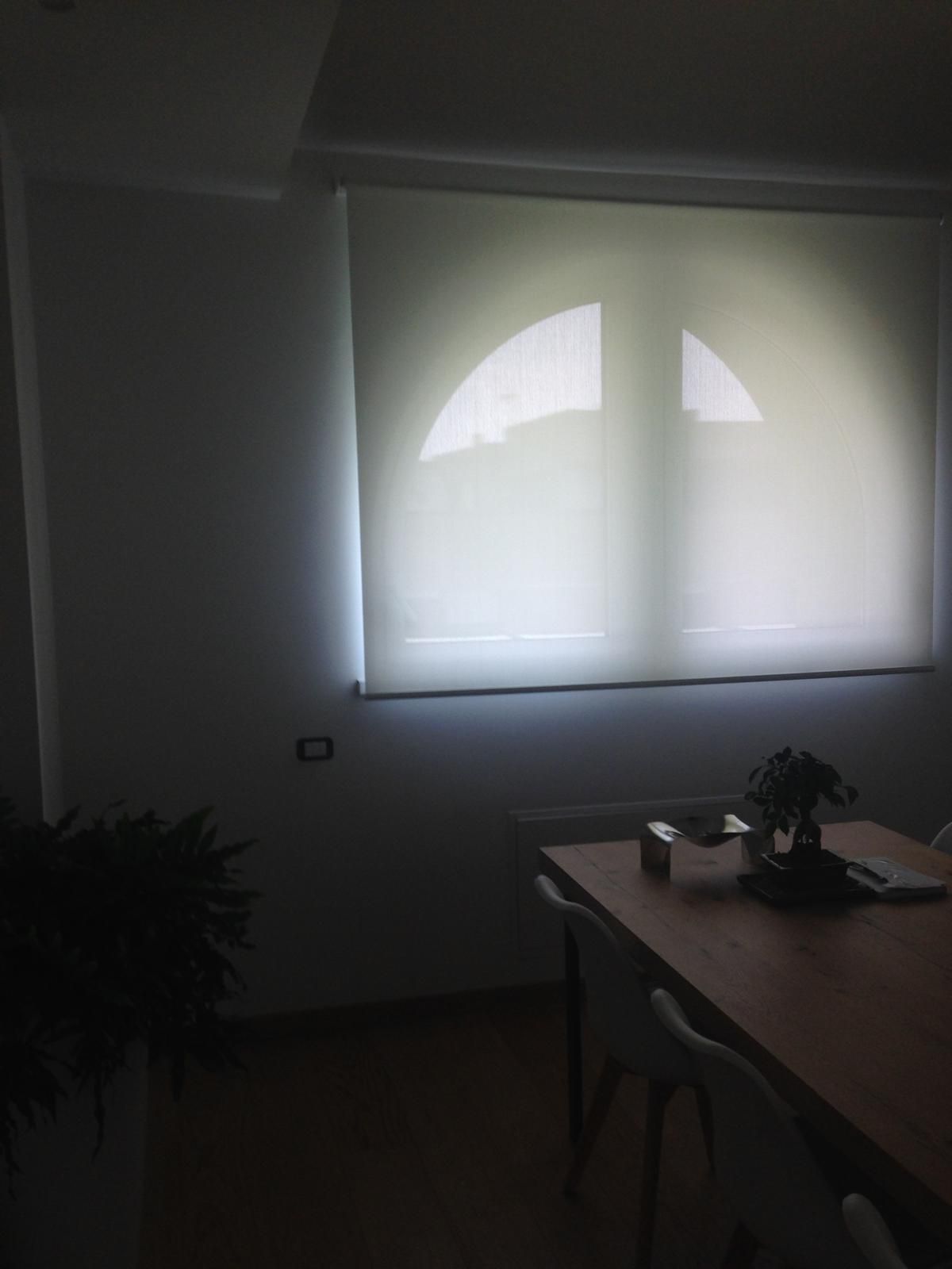 Una finestra luminosa con una tenda a rullo bianca illumina un tavolo da pranzo con sedie e una pianta in una stanza poco illuminata.