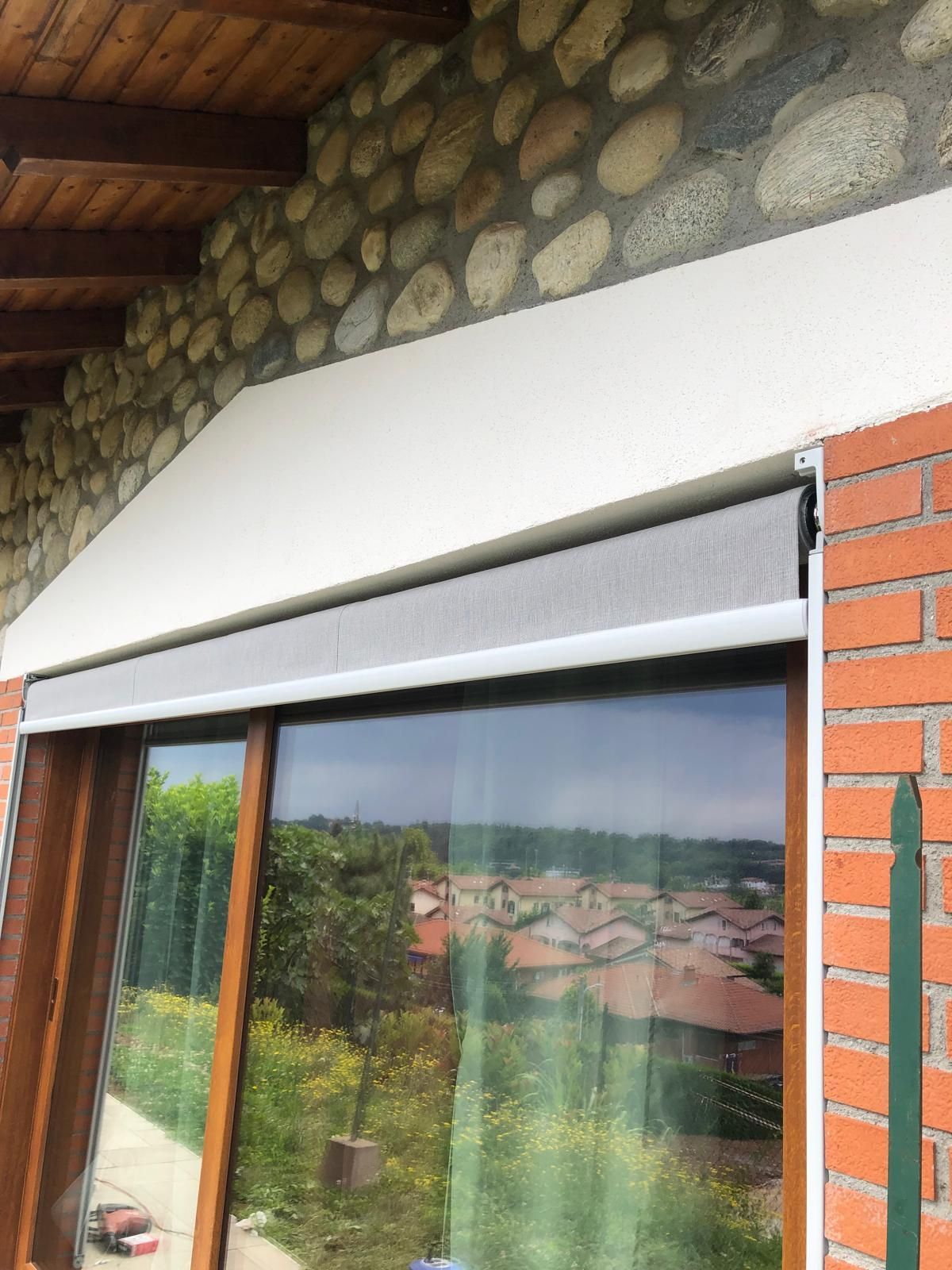 Una tenda a rullo installata sopra un'ampia finestra con cornice in legno, posizionata sotto una tenda da sole bianca inclinata su un muro di pietra.