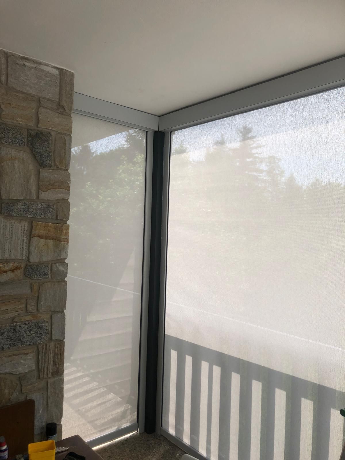 Un muro di pietra delimita una veranda chiusa da zanzariere, le cui finestre sono coperte da tende a rullo di colore chiaro che si affacciano su una foresta.