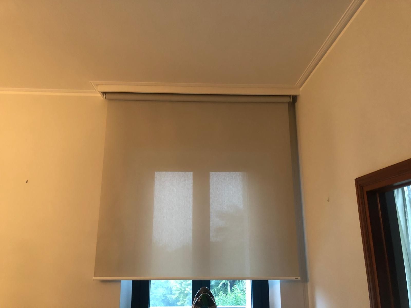 In una stanza con pareti color crema, sopra una finestra è montata una tenda a rullo grigio chiaro.