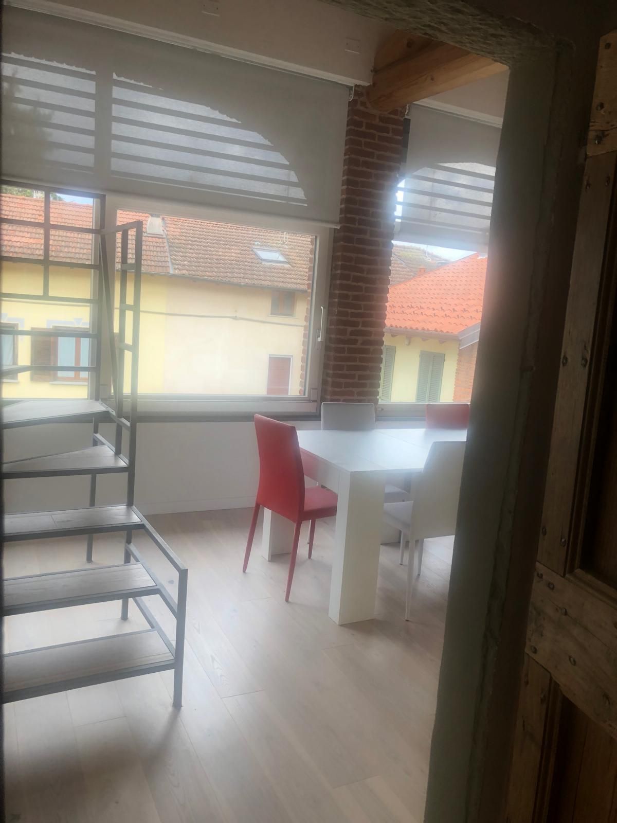 Una luminosa sala da pranzo, con un tavolo bianco e sedie bianche e rosse, si intravede attraverso una porta vicino a una scala in metallo.