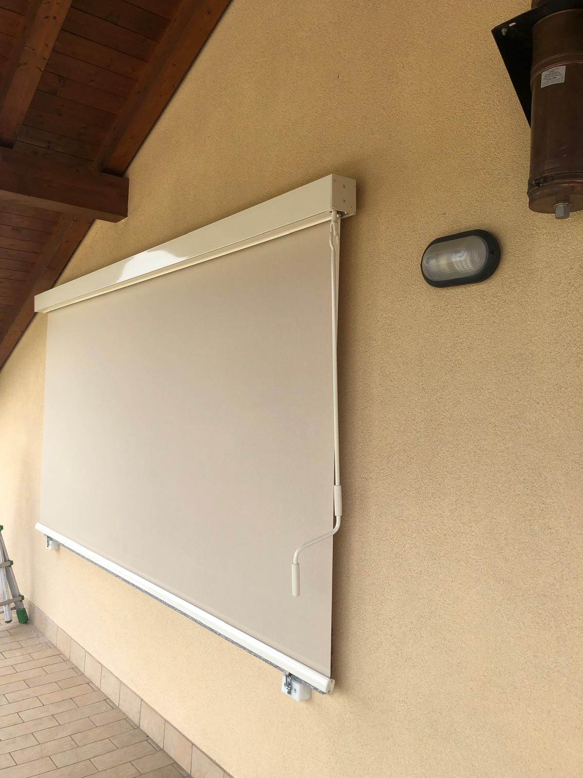Tenda a rullo beige per esterni, montata su una parete in stucco color sabbia, accanto a un'applique e a una grondaia in legno.