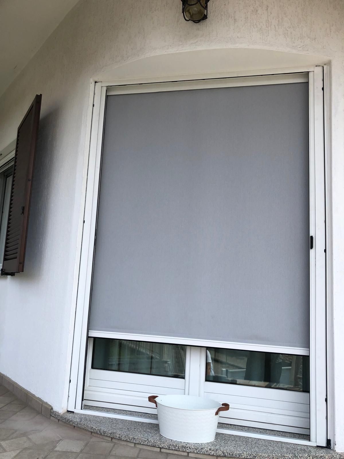 Una tenda a rullo grigia da esterno copre parzialmente una finestra bianca su un balcone, sul quale si trova un piccolo secchio bianco.