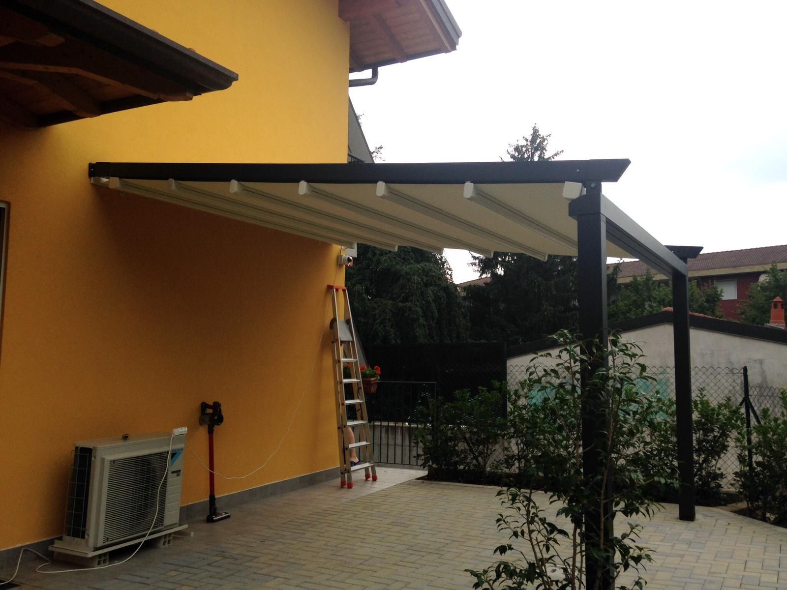Una pergola retrattile in tessuto fissata alla parete esterna di una casa gialla, sopra un patio pavimentato.