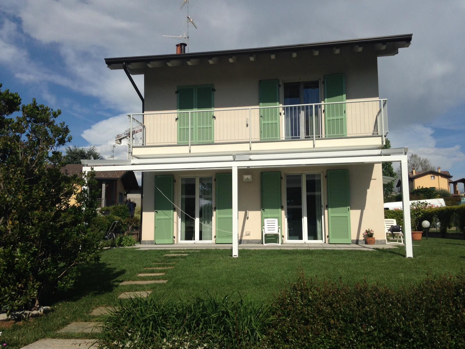 Una casa a due piani di colore chiaro con persiane verde pallido, un balcone, un pergolato bianco e un prato.
